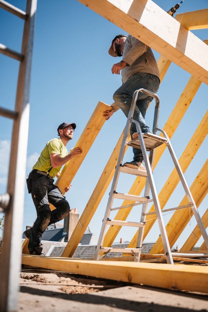 pexels-photo-31763537 Two construction workers building a wooden roof structure on a sunny day.