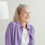 pexels-photo-8307321-8307321-1 A cheerful senior woman with grey hair wearing a purple shirt smiles indoors.